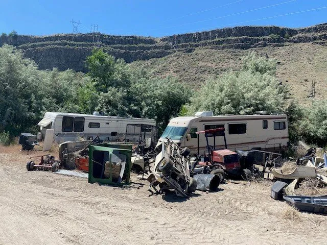 An outdoor scene with old, abandoned vehicles and assorted debris scattered around a dirt area.